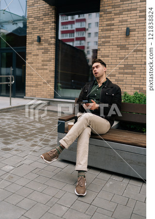 Full length vertical portrait of young man with grimace on face reacting to stupid message on phone sitting on bench, holding smartphone, with coffee cup next to him, enjoying fresh air. 121838824