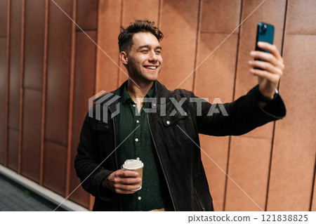 Cheerful man making a video call with smartphone while holding coffee cup, standing on background of modern building facade in urban environment. Concept of modern communication and urban lifestyle. Cheerful man making a video call with smartphone while holding coffee cup, standing on background of modern building facade in urban environment. Concept of modern communication and urban lifestyle. 121838825
