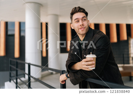 Portrait of handsome young man enjoying coffee break, leaning against railing outside modern office building, smiling looking away at beautiful cityscape. Concept of modern successful city lifestyle. 121838840