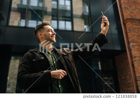 Low-angle view of cheerful handsome man holding smartphone in hand making video call, standing in front of modern buildings in city center, looking to screen with smile expression. Low-angle view of cheerful handsome man holding smartphone in hand making video call, standing in front of modern buildings in city center, looking to screen with smile expression. 121838858