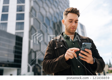 Low-angle portrait of focused successful business man using smartphone outside modern office center, confidently engaging in business communication on mobile app, looking to screen outdoors. Low-angle portrait of focused successful business man using smartphone outside modern office center, confidently engaging in business communication on mobile app, looking to screen outdoors. 121838859