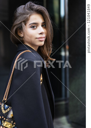 Portrait young woman wearing stylish Black Clothing With Small Handbag, beautiful model stands, poses in urban city. Modern high fashion, contemporary street style on Dark Background Portrait young woman wearing stylish Black Clothing With Small Handbag, beautiful model stands, poses in urban city. Modern high fashion, contemporary street style on Dark Background 121841849
