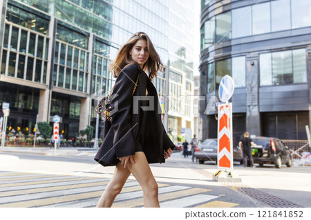 Stylish Young Fashion Woman With Long Hair Walking Across Pedestrian Crossing In City in Front of Modern Building, Wearing Black Mini Dress And blazer, Trendy Urban Street Style Model 121841852
