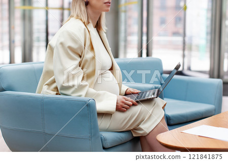 Close up Pregnant woman in a white business suit with a blazer working on a laptop in an office, indoors, sitting in a blue chair. Professional expecting mother multitasking at work with dedication Close up Pregnant woman in a white business suit with a blazer working on a laptop in an office, indoors, sitting in a blue chair. Professional expecting mother multitasking at work with dedication 121841875