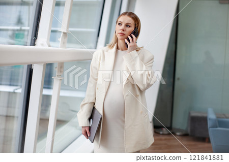 Portrait of a beautiful pregnant businesswoman in a white suit, talking on a mobile phone with a laptop in a bright office against a white and blue background. Modern motherhood into the corporate Portrait of a beautiful pregnant businesswoman in a white suit, talking on a mobile phone with a laptop in a bright office against a white and blue background. Modern motherhood into the corporate 121841881
