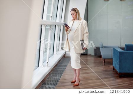 Portrait of beautiful pregnant businesswoman confidently working on laptop in a modern business office with a light blue background, wearing a stylish suit and blazer, representing female empowerment Portrait of beautiful pregnant businesswoman confidently working on laptop in a modern business office with a light blue background, wearing a stylish suit and blazer, representing female empowerment 121841900