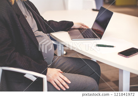 Close up Pregnant woman working on laptop at white desktop. Cropped pregnant businesswoman typing on laptop while sitting at her working place in office 121841972