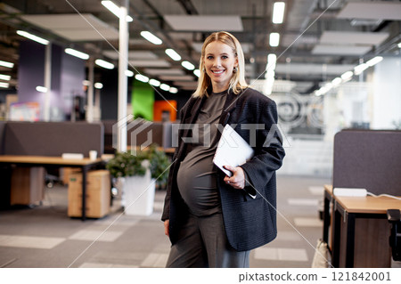 Confident pregnant businesswoman standing in a modern office, holding a tablet and smiling, showcasing balance between career and motherhood in a professional environment 121842001