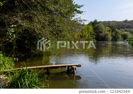 Calm riverbank with wooden dock surrounded by lush greenery on a sunny day 121842564
