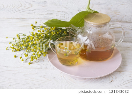 Herbal medicinal tea with chamomile flowers in a glass teapot, a bouquet of fresh flowers, on a wooden light background. 121843082