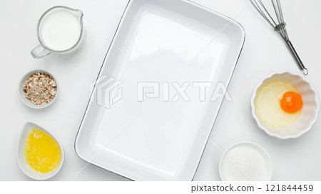 Flat lay composition for cooking preparation. Empty white baking dish with ingredients for baking, including egg, milk, butter, sugar, and nuts Flat lay composition for cooking preparation. Empty white baking dish with ingredients for baking, including egg, milk, butter, sugar, and nuts 121844459
