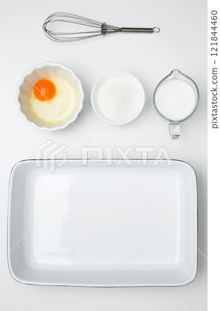 Flat lay composition for cooking preparation. Empty white baking dish with ingredients for baking, including egg, milk, butter, sugar, nuts 121844460