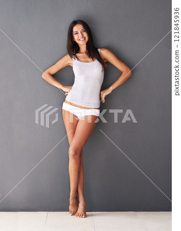 Happy, carefree and attractive woman in her underwear standing against a grey wall at home. Relaxed, beautiful and stunning young female in her house wearing a tank top and panties 121845936