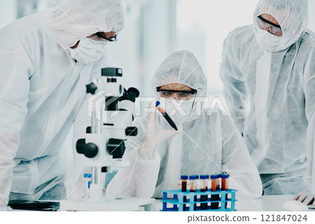 Group of scientists conducting medical research in hazmat suits and protective gloves, for coronavirus cure in a laboratory. Researchers conducting experiments and examining test tube for results. 121847024