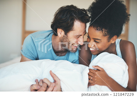 Happy, loving and funny interracial couple smiling and laughing while lying in bed and sharing a tender moment. Carefree husband and wife relaxing and having fun while bonding in the bedroom 121847229