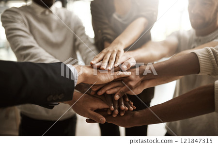 Businesspeople with their hands together in a huddle pile showing support, teamwork and close collaboration at the workplace. Closeup of a diverse group of colleagues standing in unity at the office 121847315