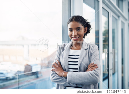 Happy, confident and professional young business woman folding her arms while standing in a modern office. Portrait of a black woman leader smiling, looking proud, powerful, ready and focused 121847443