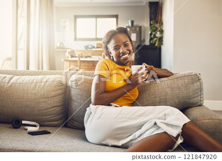 Beautiful, pretty and happy woman relaxing, drinking coffee and resting on the couch in the living room alone at home. Portrait of a smiling, cheerful and joyful black female enjoying a warm drink 121847920