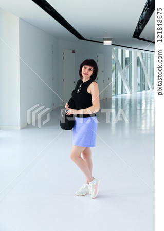 Woman in a Short Purple Skirt Standing Gracefully in a Long White Corridor, Embracing the Contrast of Colors and the Essence of Style, Capturing a Moment of Elegance and Poise in an Urban Environment 121848675