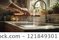 Hand washing a wooden cutting board under running water in a kitchen sink. Bright kitchen with plants in the background 121849301