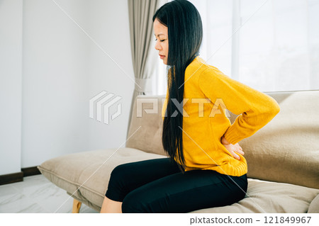 Depicting unbearable back pain, Asian woman seated on a sofa holds her lower back. Illustrating chronic backache discomfort and the necessity of medical care for back problems. health care concept 121849967