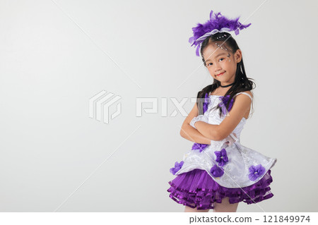 Cheerleader girl in a white and purple dress with a layered skirt, purple feather headpiece, and crossed arms. Studio photo with a clean white backdrop, showcasing her confident pose 121849974