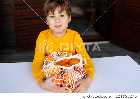 A cheerful child in a vibrant yellow sweater hugs a mesh bag brimming with citrus fruits while seated at a table. A cheerful child in a vibrant yellow sweater hugs a mesh bag brimming with citrus fruits while seated at a table. 121850279