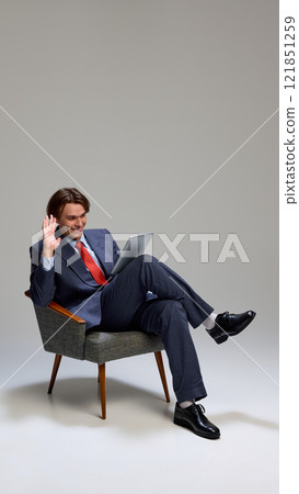 Man sitting in chair, dressed suit with red tie, holding tablet and waving hand with smile against gray studio background. Man sitting in chair, dressed suit with red tie, holding tablet and waving hand with smile against gray studio background. 121851259