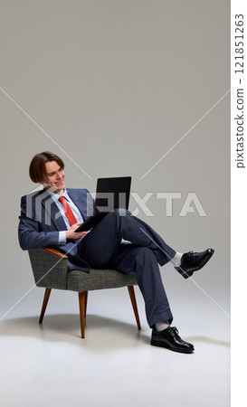 Man sitting in chair, dressed suit with red tie, holding black laptop and laughing in relaxed pose against gray studio background. Man sitting in chair, dressed suit with red tie, holding black laptop and laughing in relaxed pose against gray studio background. 121851263