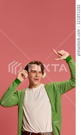 Man enjoying music with headphones, gesturing playfully with hand, wearing green cardigan and white t-shirt against soft pink studio background. Man enjoying music with headphones, gesturing playfully with hand, wearing green cardigan and white t-shirt against soft pink studio background. 121851282