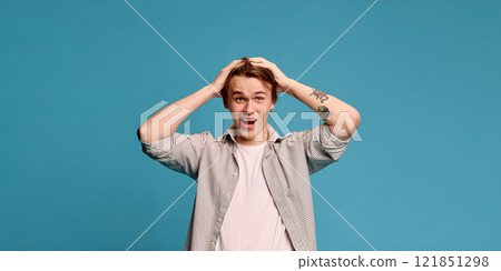 Man holding head with hands, expressing excitement, dressed in plaid shirt and casual jeans against vibrant blue studio background. 121851298