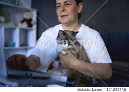 a senior woman works from home on a laptop with a pet, a cat 121852536