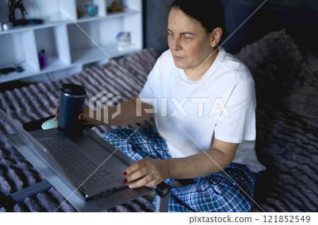 a senior woman works from home on a laptop and drinks coffee from a thermos mug a senior woman works from home on a laptop and drinks coffee from a thermos mug 121852549