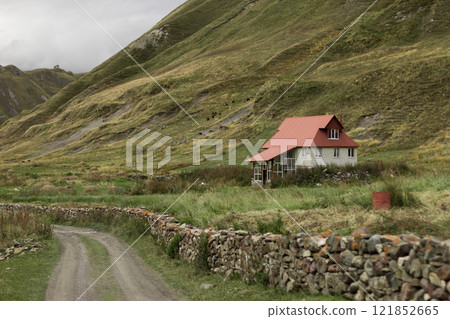 A charming house with a red roof rests by a winding dirt road in the Georgian autumn hills, surrounded by rolling slopes and a cloudy daytime sky A charming house with a red roof rests by a winding dirt road in the Georgian autumn hills, surrounded by rolling slopes and a cloudy daytime sky 121852665