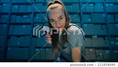 Positive Female Warehouse Worker Looking at Camera, Scanning Cardboard Box Using Scanner 121854040