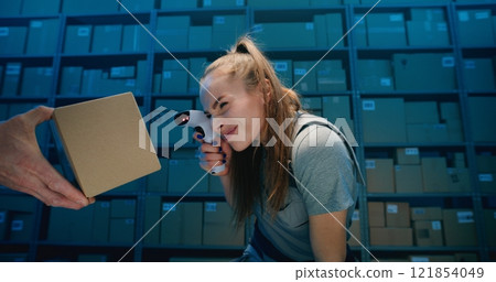 Positive Female Logistics Specialist Looking at Camera, Scanning Cardboard Box with Scanner in Warehouse 121854049