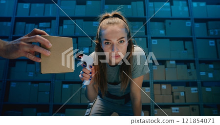 Tired Female Warehouse Employee Looking at Camera, Scanning Cardboard Boxes Using Scanner 121854071
