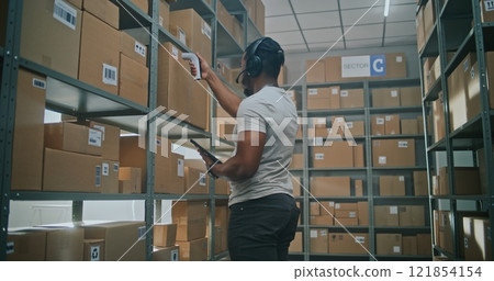 Worker Wearing Robotic Exoskeleton Puts Cardboard Box on Shelf. Logistics Specialist Scans Parcel Worker Wearing Robotic Exoskeleton Puts Cardboard Box on Shelf. Logistics Specialist Scans Parcel 121854154