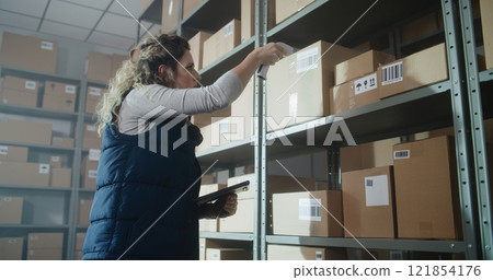 Female Warehouse Associate Scans Cardboard Boxes, Checks Inventory Using Tablet Computer Female Warehouse Associate Scans Cardboard Boxes, Checks Inventory Using Tablet Computer 121854176