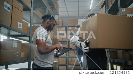 African American Manager Scans Parcels, Worker in Exoskeleton Walks with Cardboard Box African American Manager Scans Parcels, Worker in Exoskeleton Walks with Cardboard Box 121854185