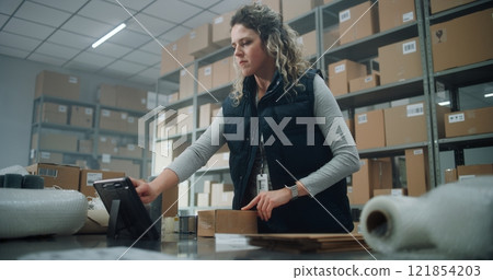 Female Warehouse Clerk Packs Cardboard Parcel for Shipping, Uses Tablet Computer 121854203