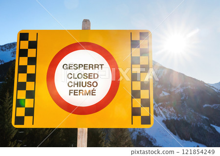 Close-up yellow multilingual warning sign Closed on snowy mountain resort under clear blue skies against sunny Alps landscape. Safety in winter mountain sports concept. Avalanche weather risk danger 121854249