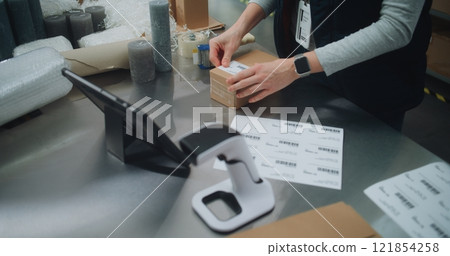 Female Postal Worker Packs Cardboard Parcel, Prepares for Delivery, Uses Tablet Computer 121854258
