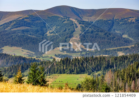 Majestic mountain landscape with lush forests and distant ski trails under clear blue sky Majestic mountain landscape with lush forests and distant ski trails under clear blue sky 121854409