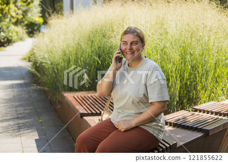 30s cheerful woman dressed in casual look sitting on the bench and talking on smartphone on the city street. Female relaxing outdoors. Urban concept. 121855622