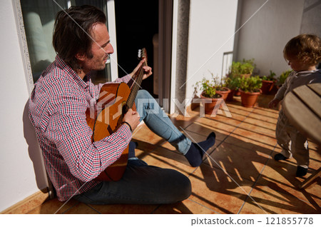 Man Playing Acoustic Guitar on Terrace While Child Observes Interaction 121855778