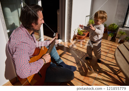 Father playing guitar for young child on a sunny balcony 121855779