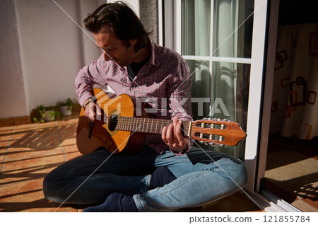 Man playing acoustic guitar while sitting on a sunny home patio Man playing acoustic guitar while sitting on a sunny home patio 121855784