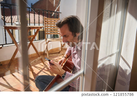 Man sitting on patio playing guitar and checking smartphone Man sitting on patio playing guitar and checking smartphone 121855786