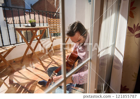 Man playing guitar on a sunny balcony with wooden furniture and glass door reflection Man playing guitar on a sunny balcony with wooden furniture and glass door reflection 121855787